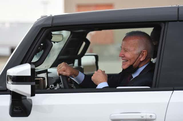 Joe Biden sits at the wheel of a electric vehicle, smiling