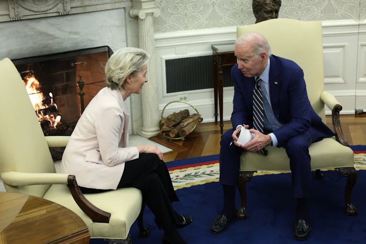 Biden and von der Lyden talk in the Oval Office. They're leaning foward toward each other in their chairs and smiling.