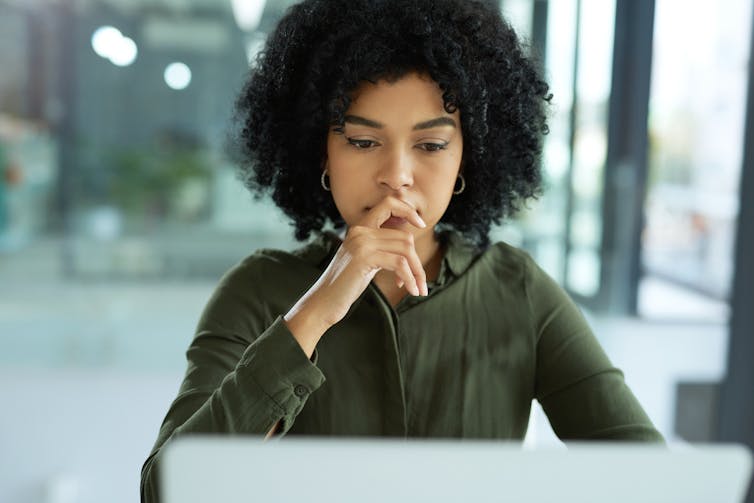 A woman with curly black hair sits looking thoughtful as she reads something on a laptop.