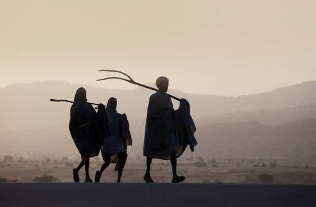 Three people walking up a rural road, carrying farm implements.