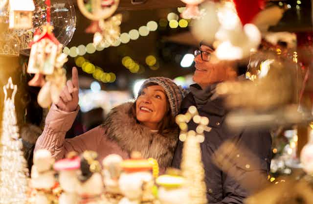 A middle aged couple smiles and points at items in a Christmas display, they are surrounded by out of focus glowing decorations
