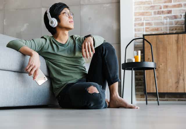 A young asian man wearing large over ear headphones, sat on the floor. 