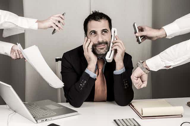 Un hombre habla por teléfono mientras otras cuatro manos le alcanzan más teléfonos, papeles, relojes, etc.