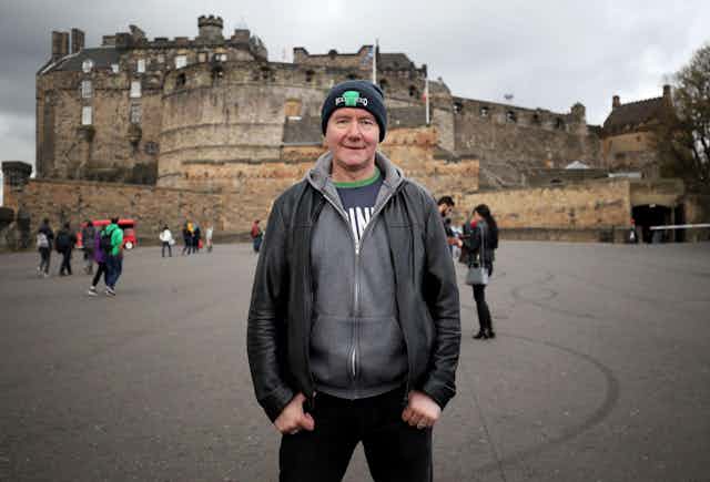 Irvine Welsh stands in Edinburgh's Royal Mile wearing a hat and hoodie.
