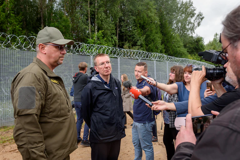 Edgars Rinkevics répond à des journalistes devant un grand grillage, aux côtés d'un homme en uniforme