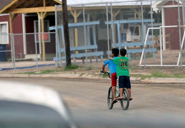 Two boys a bicycle ride pas a school with a dirt fenced-in playground 