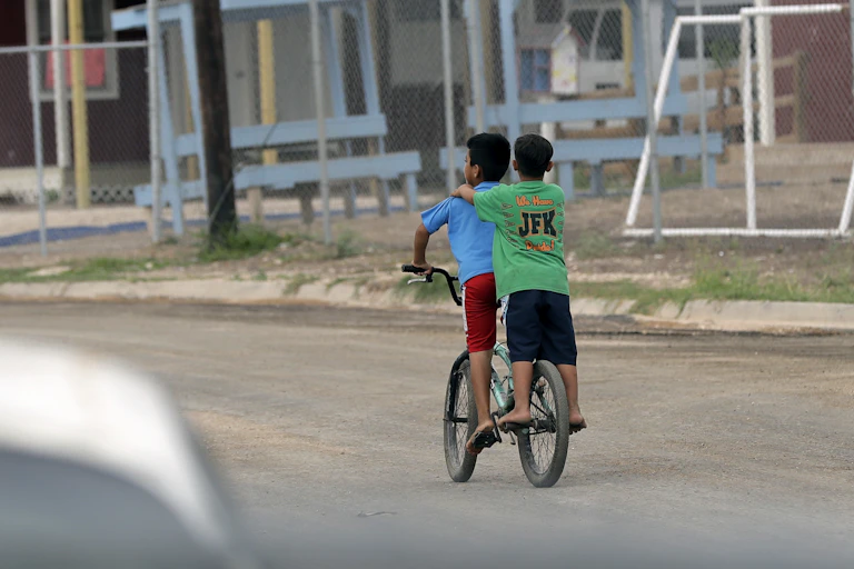 Two boys a bicycle ride pas a school with a dirt fenced-in playground