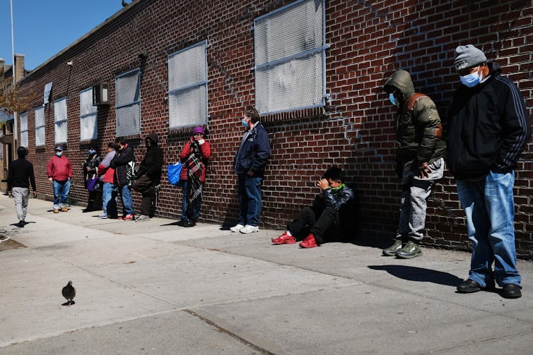 People wearing face masks lean against a brick wall as they wait their turn to enter the food bank building.