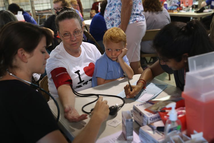 A young child, looking bored, sits on a woman's lap as a nurse tests her blood pressure.