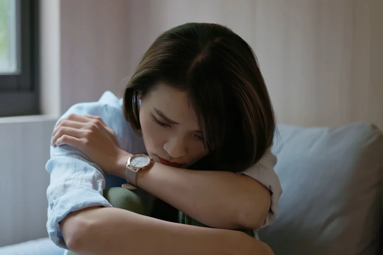 A young Chinese woman with a sad expression on her face, sitting on a couch and holding her knees to her chest