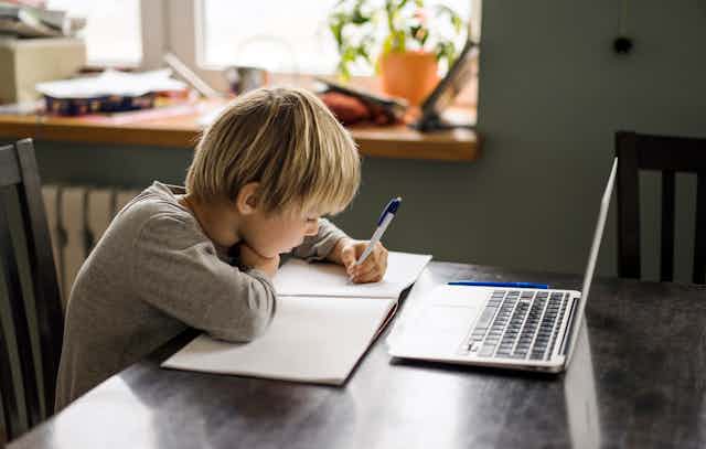 Duduk di sebuah meja, dengan laptop di depannya, seorang anak laki-laki menulis di buku catatannya.