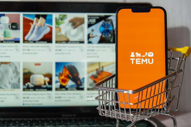 Miniature shopping trolley containing phone with Temu logo on screen, in front of laptop showing products for sale