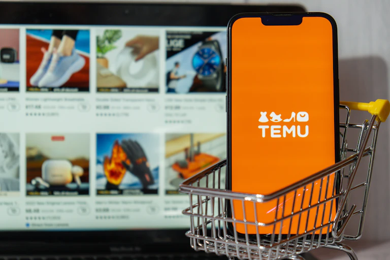 Miniature shopping trolley containing phone with Temu logo on screen, in front of laptop showing products for sale