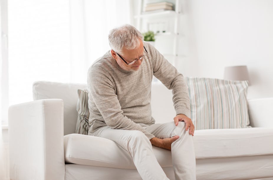 An older man sitting on a couch holds his knee in pain.