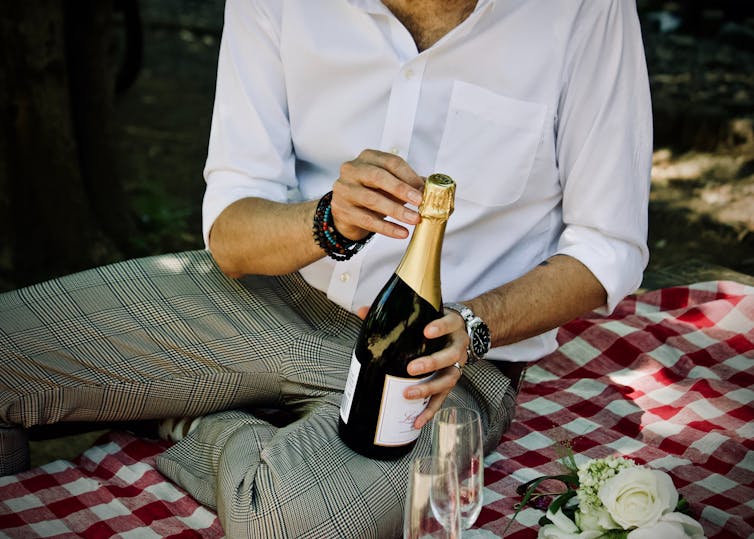 A man sits on a picnic blanket and opens a bottle of champagne.