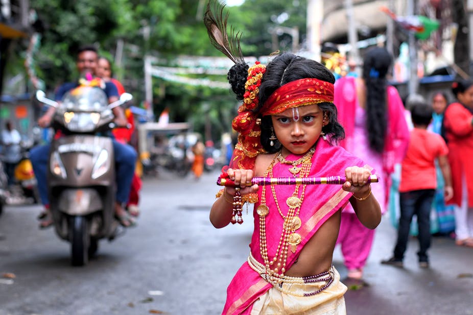 A child in colorful clothing holding a flute and wearing a peacock feather in his hair.