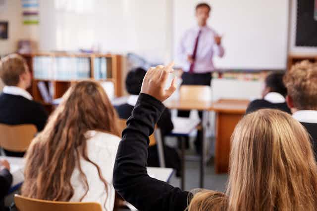 View from behind of a female student raising her hand in a classroom, as the teacher in the background lectures to the class