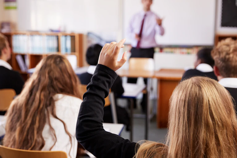 View from behind of a female student raising her hand in a classroom, as the teacher in the background lectures to the class