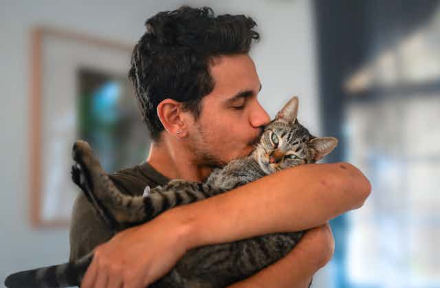Un jeune homme aux cheveux noirs tenant et embrassant un chat tabby