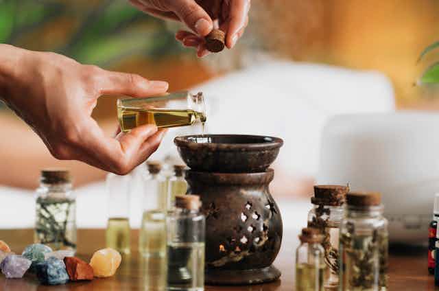 A hand pouring a small jar of oil into a diffuser, with other jars of oils, herbs and crystals sitting nearby. 