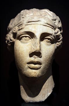 A statue of the head of a woman with curly hair, whose nose is partially missing, against a black background.