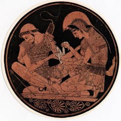 A circular, red and black image of two men in battle gear as one binds the other's wound.