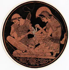 A circular, red and black image of two men in battle gear as one binds the other's wound.