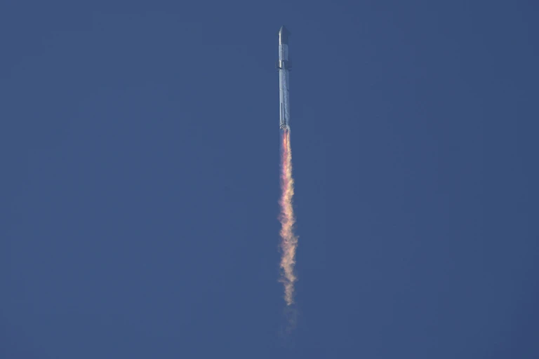 A rocket blasting off, with orange flame coming out of the end of the rocket, all against a blue sky.