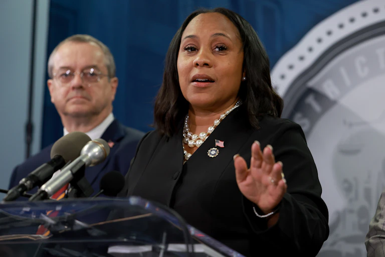 A woman wearing an American flag pin on her black suit jacket and white pearls around her neck, holds her hand her left hand at breast level, fingers pointed forward, as she speaks from behind a lectern.