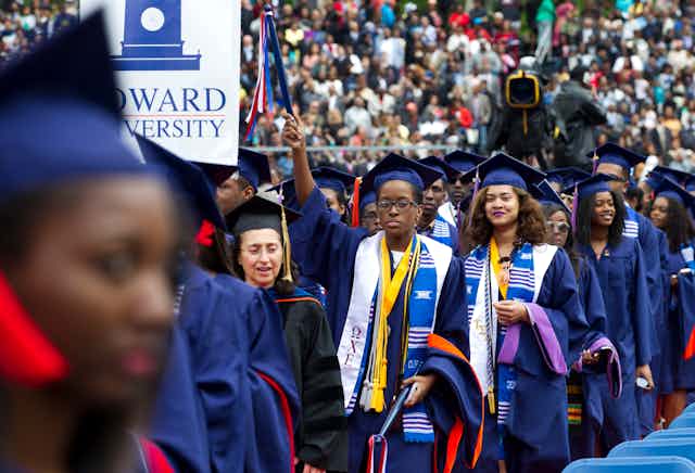 Studens wearing blue caps and gowns assemble at a graduation ceremony.