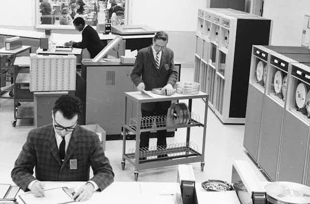 Black and white photo of antiquated computer equipment and three workers in office attire