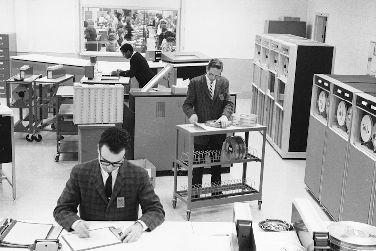 Black and white photo of antiquated computer equipment and three workers in office attire