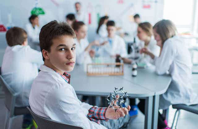 A teenage boy holds an artificial model of a molecule in a laboratory and looking at the camera, while his classmates are in the background.