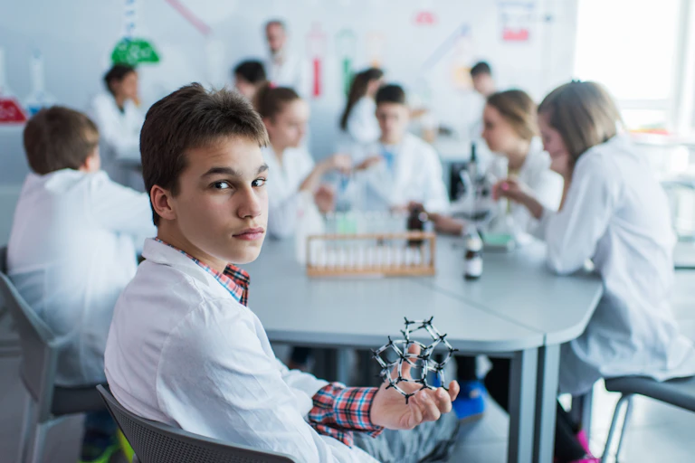 A teenage boy holds an artificial model of a molecule in a laboratory and looking at the camera, while his classmates are in the background.