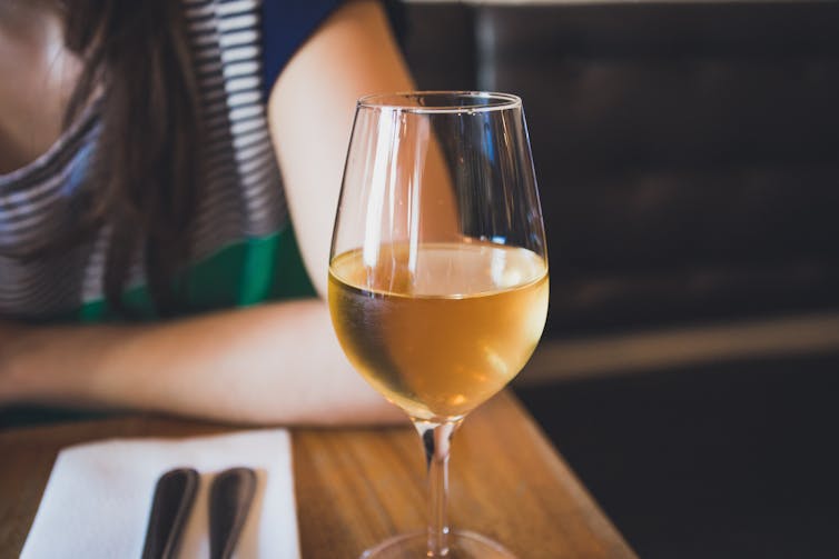 A glass of white wine sits in front of a woman at a table in a restaurant.