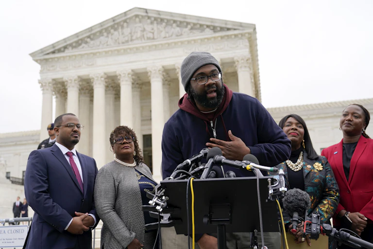 A man with a beard and glasses, wearing a gray hat, speaks in front of a large, white building.