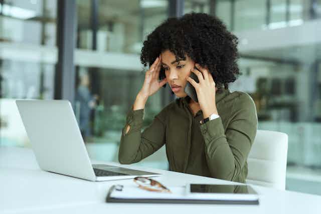 Woman at office in front of laptop holds cellphone to ear