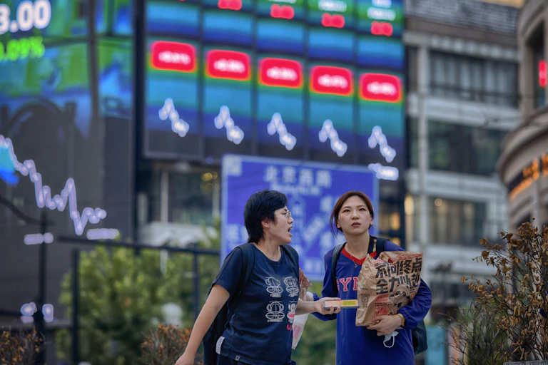 Two women stand in from of screens showing stock prices