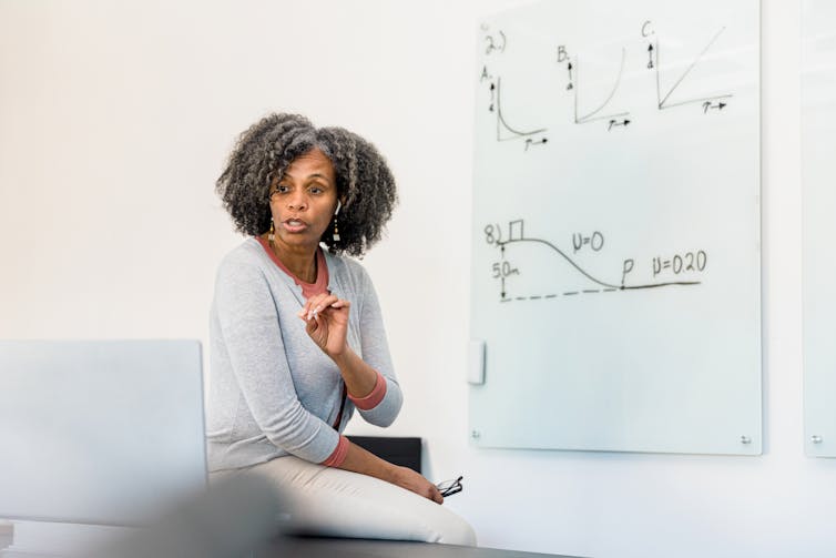 A woman sits on her desk near a whiteboard with an equation written in black marker.