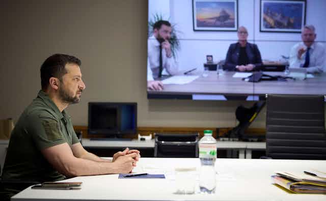 Side profile of serious man with beard in green shirt with a video screen in the background showing three men, two in shirt and tie.
