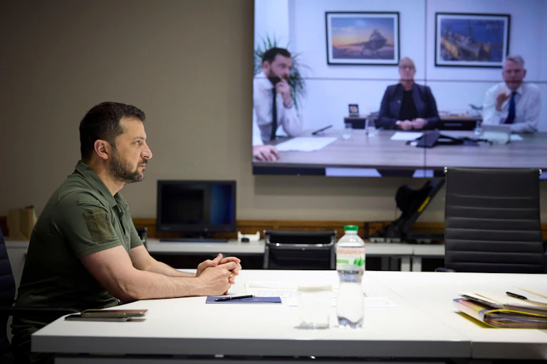 Side profile of serious man with beard in green shirt with a video screen in the background showing three men, two in shirt and tie.