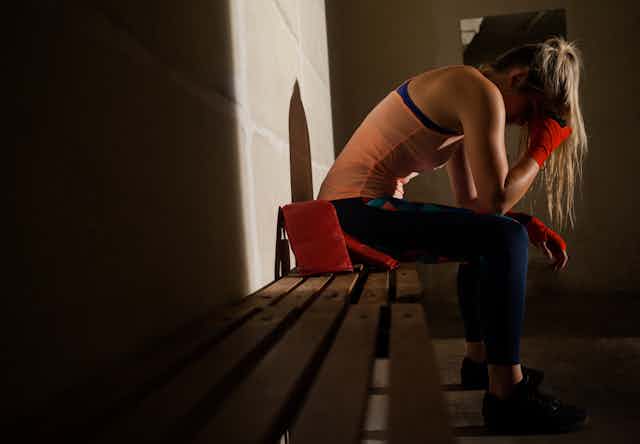 Side view of a young woman in athletic wear sits sadly on a bench in a dark locker room, with her head in her hands