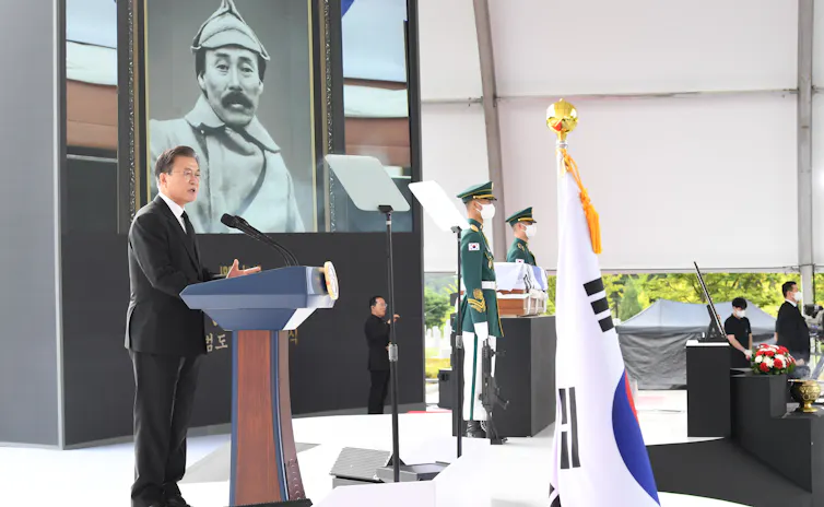 Former South Korean president Moon Jae-in stands at a lectern. IN the background is a picture of Korean independence fighter Hong Beom-do