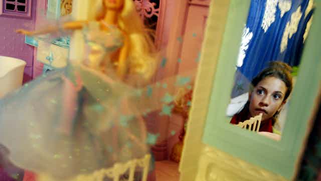 Close-up of Barbie doll on display while mirror reflects image of girl observing it.