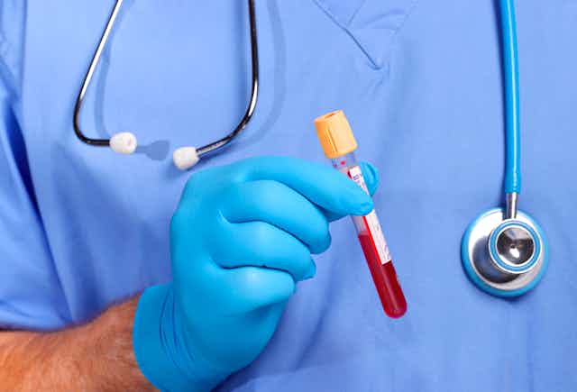 A person wearing scrubs and gloves holds up a test tube filled halfway up with blood.
