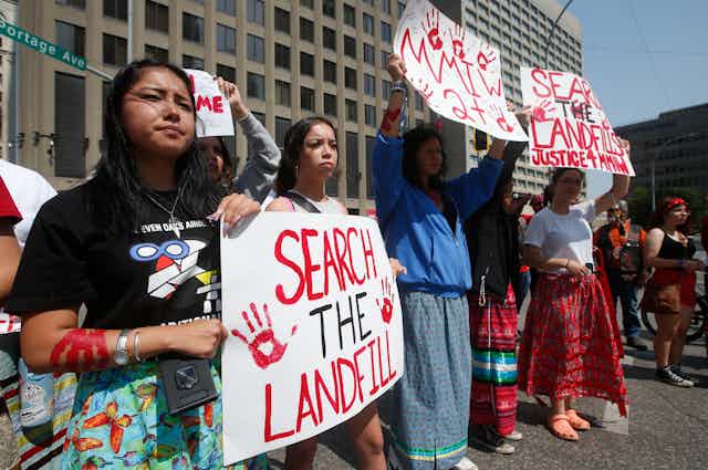 People line the streets holding posters that say 'Search the Landfill'