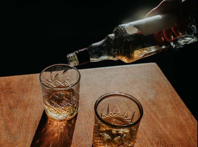 A hand pours a light brown liquor into a crystal glass, which sits next to another cyrstal glass on a wood table. 