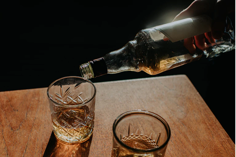A hand pours a light brown liquor into a crystal glass, which sits next to another cyrstal glass on a wood table.