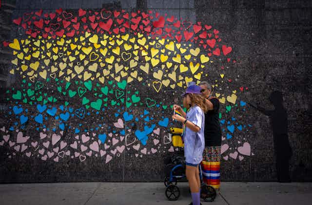 A wall with heart shapes in the colours of the rainbow flag. Two women stand beside.