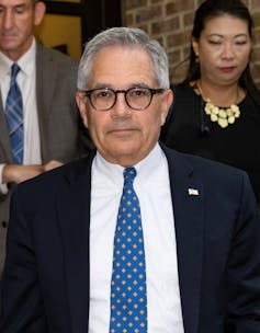 A gray-haired white man wearing a suit and a blue tie looks into the camera